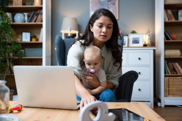 A young woman with a baby on her lap sits in front of a table with an open laptop and tablet on it