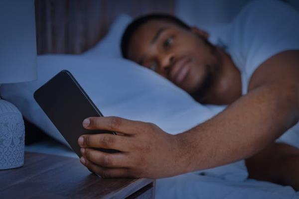 A man lying in bed on his side looks at his phone, which is resting on his bedside table